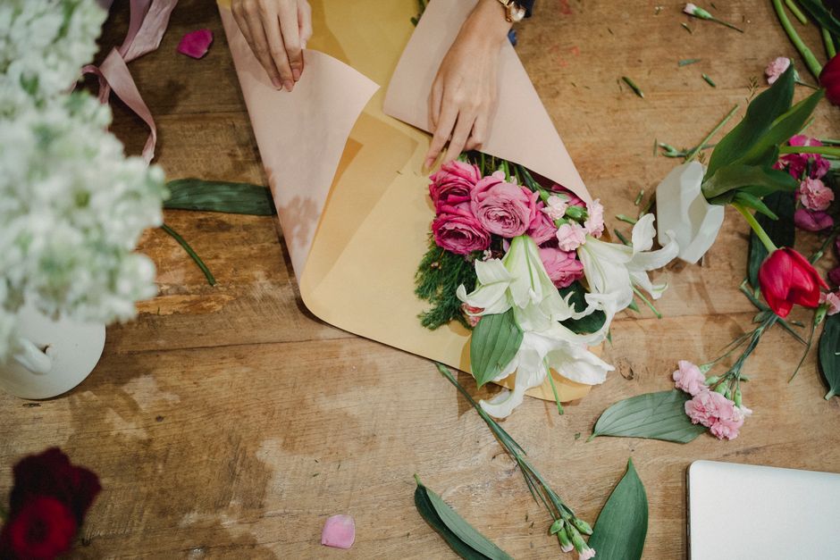 A florist at a wooden workspace arranges a floral gift, wrapping a bouquet of pink roses, white lilies, and greenery in light brown paper with a pink ribbon. The bouquet features fresh, vibrant flower