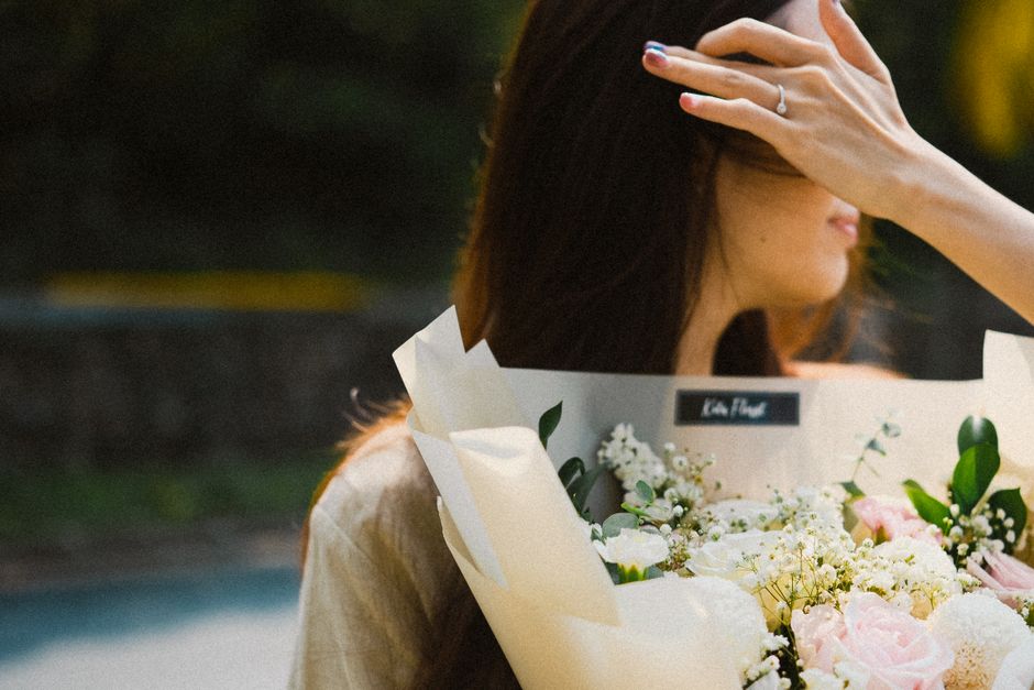 A woman with long, dark hair holding a bouquet of fresh flowers, including pale pink roses, white baby's breath, and greenery, wrapped in cream-colored paper with a black label. She is outdoors, parti