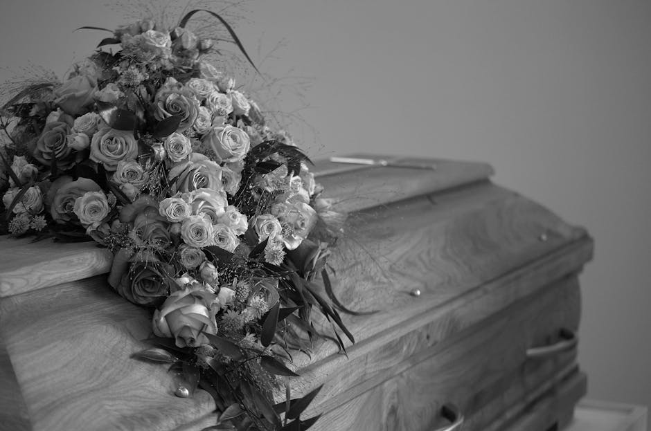 A floral arrangement resting on a wooden casket, consisting of an abundant bouquet of fresh roses and smaller blooms. The roses are in various stages of bloom, with some fully open and others partiall