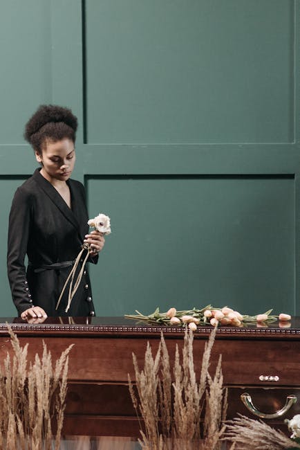 A young woman with dark, curly hair styled in a natural afro, dressed in a black blazer, stands solemnly at a wooden funeral altar. She holds a small white flower, possibly a rose or peony, in her rig