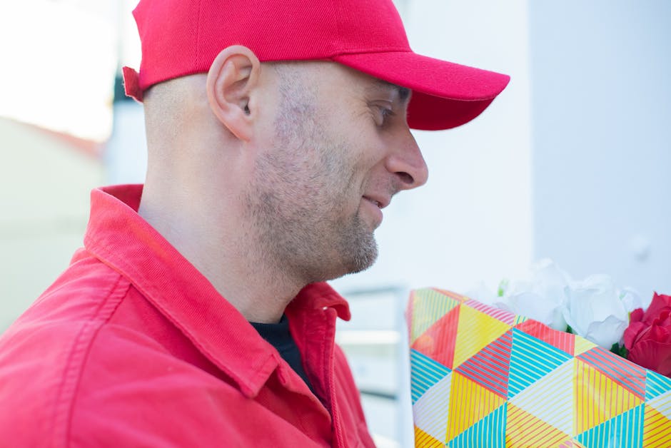 A delivery person dressed in a red uniform and matching cap is holding a floral gift wrapped in colorful geometric-patterned paper, featuring shades of yellow, blue, red, and green. The bouquet appear